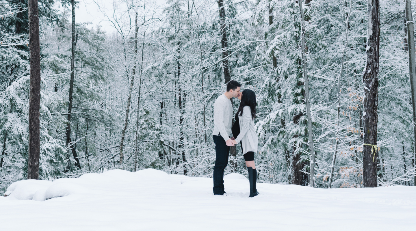 pregnant couple kissing outside in the winter in the snow with a forest behind them